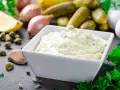 Una cremosa salsa remoulade in una ciotola bianca, circondata da ingredienti freschi come limone, capperi, aglio e cetriolini.