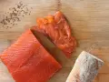 Fetta di salmone marinato, affettato e pane su tagliere di legno. Semi di cumino e sale completano la scena.
