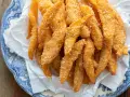 Un mucchio di bastoncini dorati di zucca fritta impanata, croccanti e salati, serviti su carta bianca in un piatto blu e bianco.