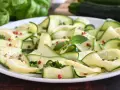 Zucchine marinate a fettine sottili, condite con erbe aromatiche e pepe rosa, su un piatto bianco.