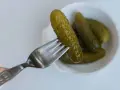 Un cetriolo sottaceto viene sollevato da una forchetta da una ciotola bianca piena di altri cetrioli sottaceto.