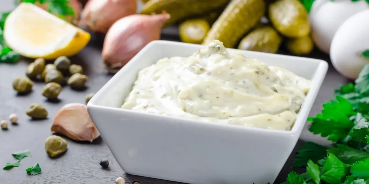 Una cremosa salsa remoulade in una ciotola bianca, circondata da ingredienti freschi come limone, capperi, aglio e cetriolini.