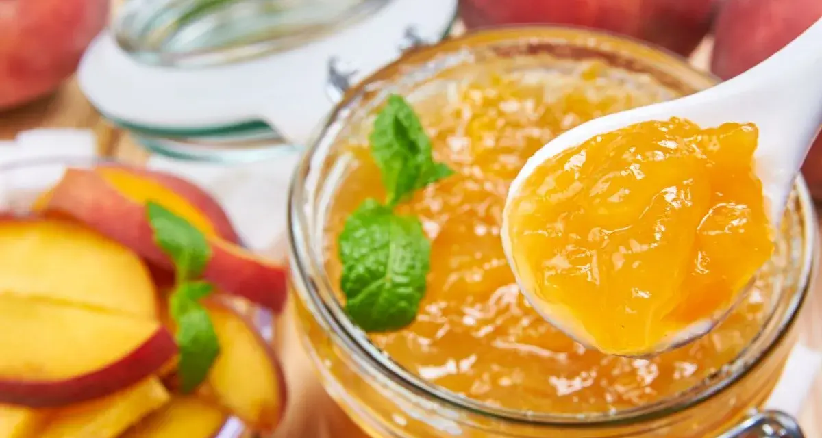 Un cucchiaio raccoglie la deliziosa marmellata di pesche da un barattolo, con pesche fresche e foglie di menta intorno.