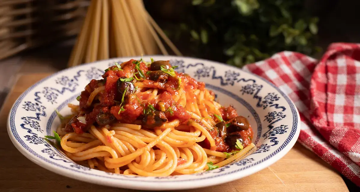 Piatto di spaghetti alla puttanesca, conditi con pomodoro, olive e capperi, guarniti con prezzemolo fresco.
