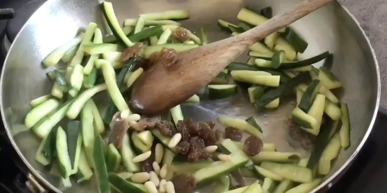 Zucchine in agrodolce veloci in padella con pinoli e uvetta, mescolate con un cucchiaio di legno.