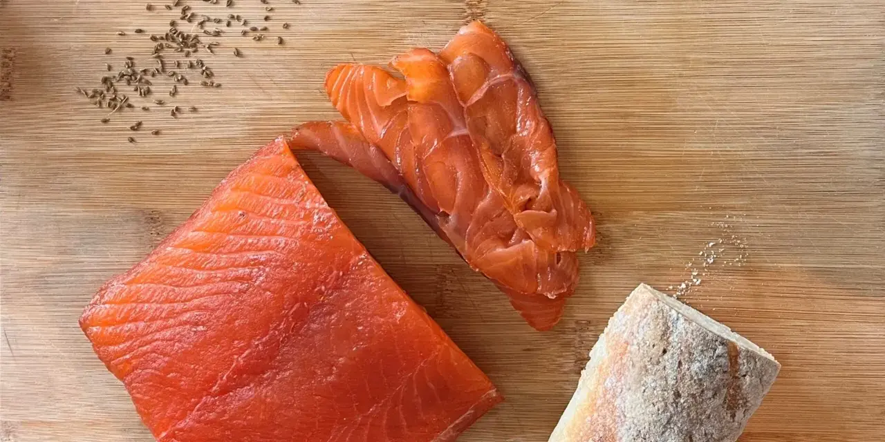 Fetta di salmone marinato, affettato e pane su tagliere di legno. Semi di cumino e sale completano la scena.