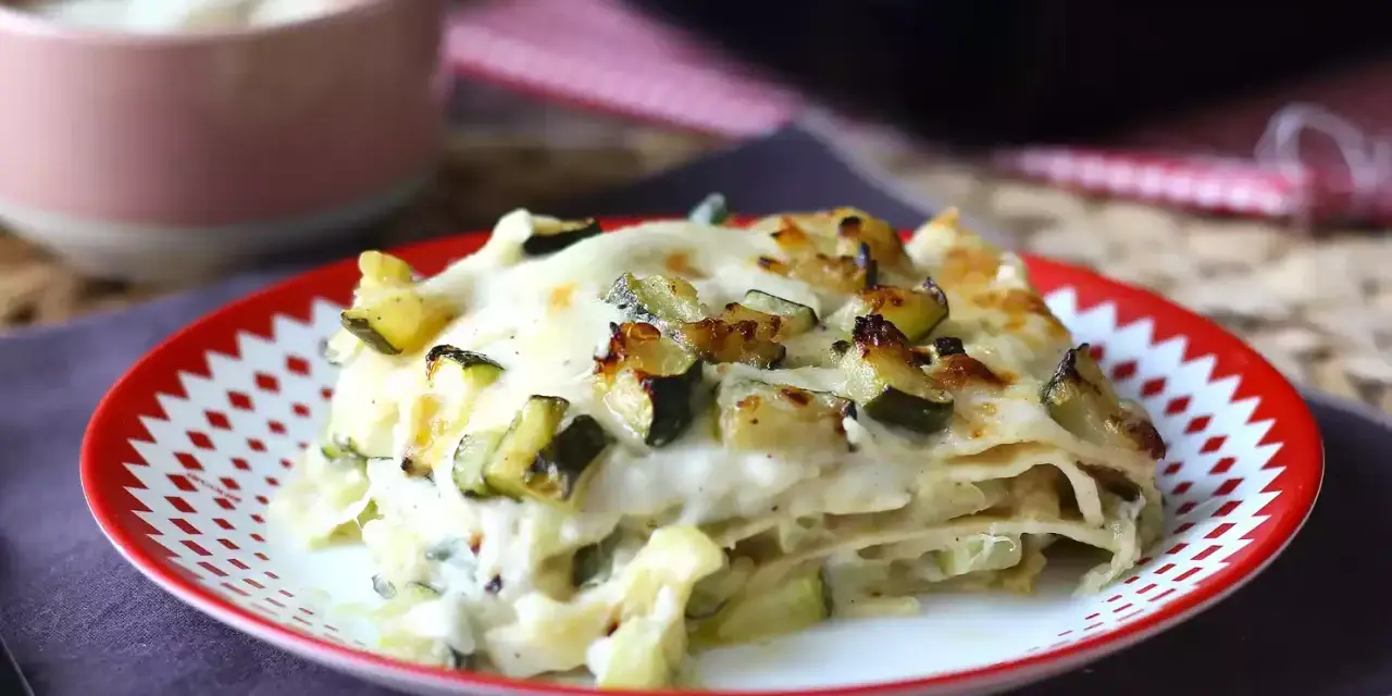Una fetta di lasagne vegetariane con zucchine e besciamella, dorata al forno, servita su un piatto rosso e bianco.