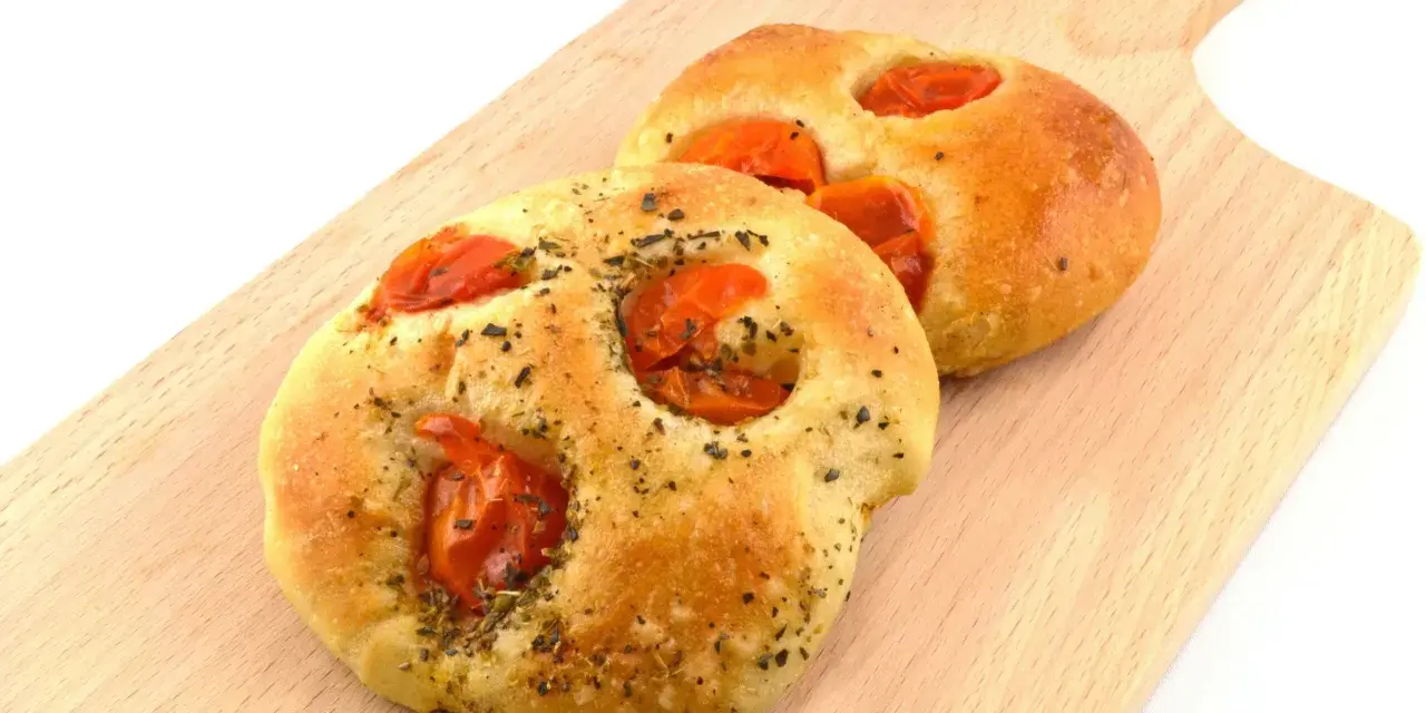 Due focaccine pugliesi dorate, condite con pomodorini e erbe aromatiche, su un tagliere di legno.