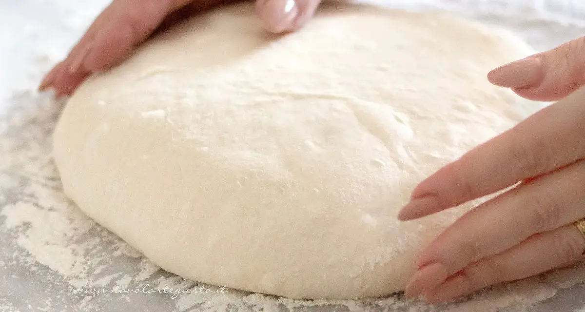Mani che lavorano un soffice impasto pizza su un piano infarinato, pronte per essere stese.