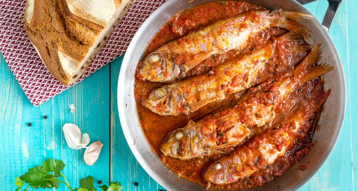 Triglie alla livornese in padella, con pane rustico, aglio e prezzemolo su tavolo blu.