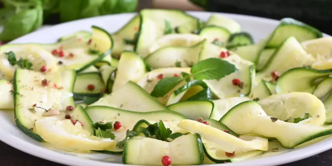 Zucchine marinate a fettine sottili, condite con erbe aromatiche e pepe rosa, su un piatto bianco.