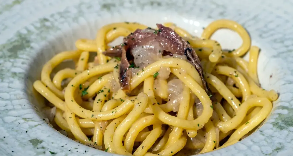 Un piatto di bigoli in salsa, con pasta spessa e condimento a base di acciughe e cipolla, guarnito con prezzemolo tritato.