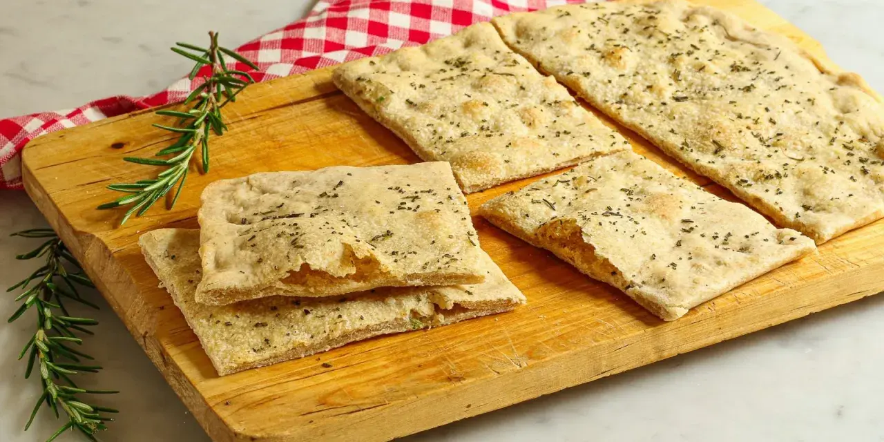 Focaccia senza lievito, croccante e profumata al rosmarino, servita su un tagliere di legno con un tovagliolo a quadri rossi.