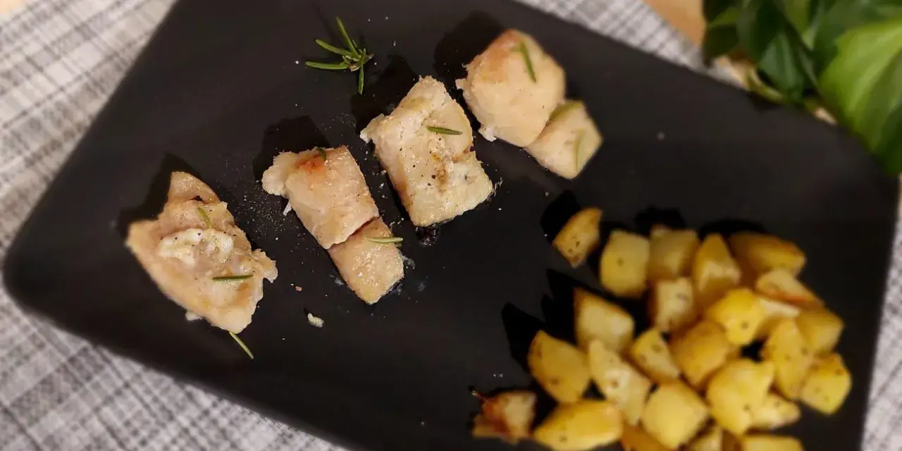 Pezzi di baccalà al forno con rosmarino e patate arrosto su piatto nero.