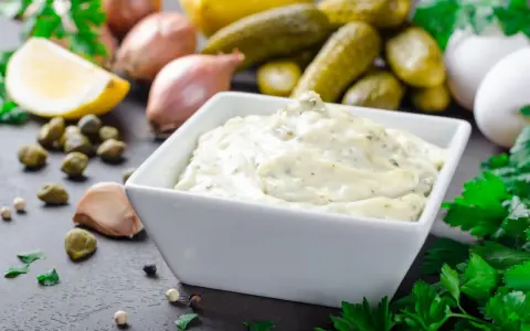 Una cremosa salsa remoulade in una ciotola bianca, circondata da ingredienti freschi come limone, capperi, aglio e cetriolini.