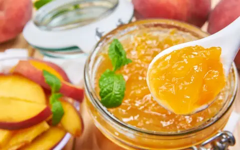 Un cucchiaio raccoglie la deliziosa marmellata di pesche da un barattolo, con pesche fresche e foglie di menta intorno.