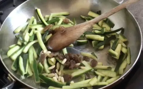 Zucchine in agrodolce veloci in padella con pinoli e uvetta, mescolate con un cucchiaio di legno.