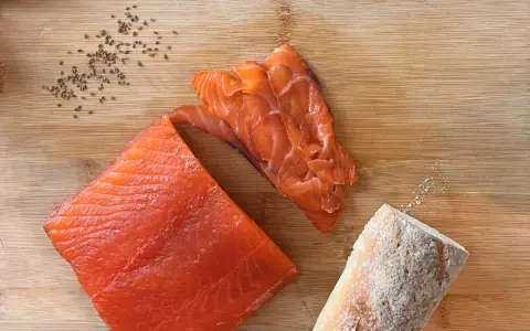 Fetta di salmone marinato, affettato e pane su tagliere di legno. Semi di cumino e sale completano la scena.