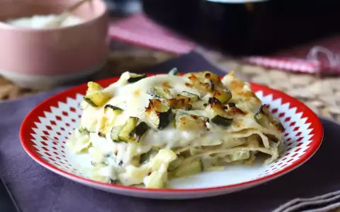 Una fetta di lasagne vegetariane con zucchine e besciamella, dorata al forno, servita su un piatto rosso e bianco.