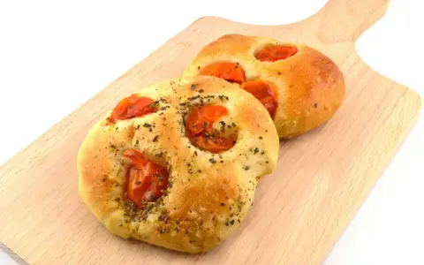 Due focaccine pugliesi dorate, condite con pomodorini e erbe aromatiche, su un tagliere di legno.