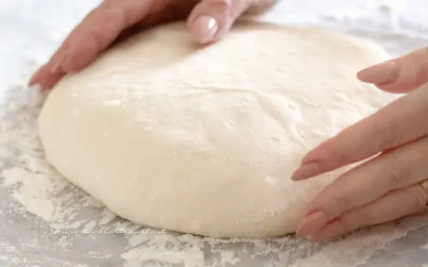 Mani che lavorano un soffice impasto pizza su un piano infarinato, pronte per essere stese.