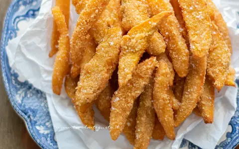 Un mucchio di bastoncini dorati di zucca fritta impanata, croccanti e salati, serviti su carta bianca in un piatto blu e bianco.