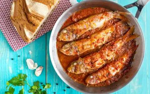 Triglie alla livornese in padella, con pane rustico, aglio e prezzemolo su tavolo blu.
