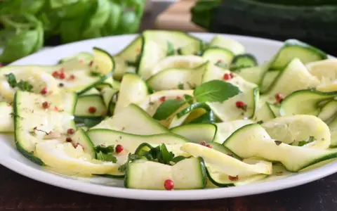 Zucchine marinate a fettine sottili, condite con erbe aromatiche e pepe rosa, su un piatto bianco.