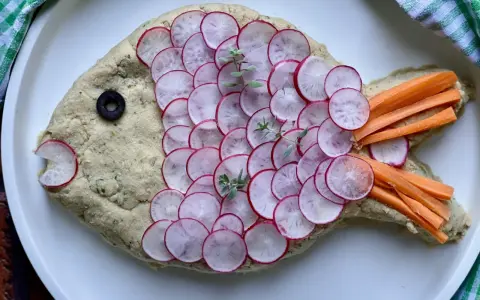 Pesce finto ricetta: un impasto cremoso decorato con ravanelli affettati, carote a bastoncino, un'oliva e erbe aromatiche.