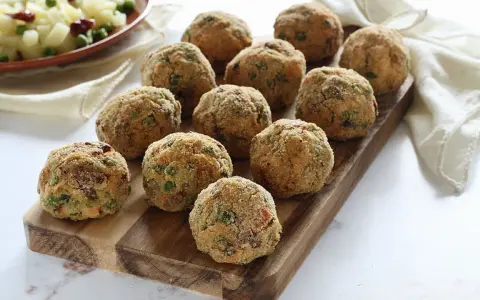 Deliziose polpette di verdure dorate e croccanti, disposte su un tagliere di legno. Un contorno di patate e piselli sullo sfondo.