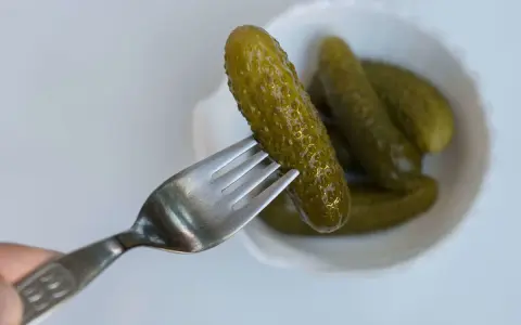 Un cetriolo sottaceto viene sollevato da una forchetta da una ciotola bianca piena di altri cetrioli sottaceto.
