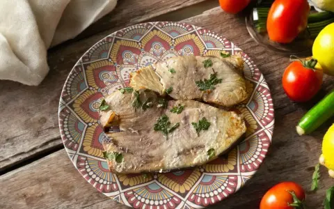 Due fette di pesce spada cotto in friggitrice ad aria, guarnite con prezzemolo, su un piatto decorato. Accanto, pomodori, limoni e zucchine.