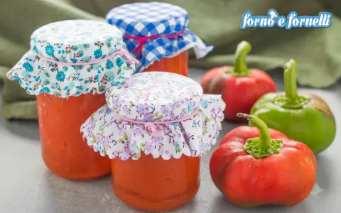 Barattoli di salsa di peperoni fatti in casa, decorati con stoffe floreali e a quadretti, accanto a peperoni freschi.