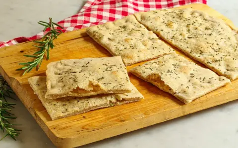 Focaccia senza lievito, croccante e profumata al rosmarino, servita su un tagliere di legno con un tovagliolo a quadri rossi.