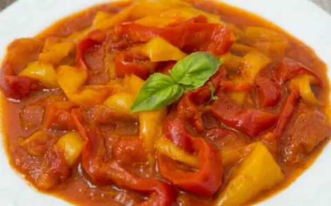 Un piatto di peperonata ricetta, con peperoni rossi e gialli cotti in salsa, guarnito con basilico fresco.