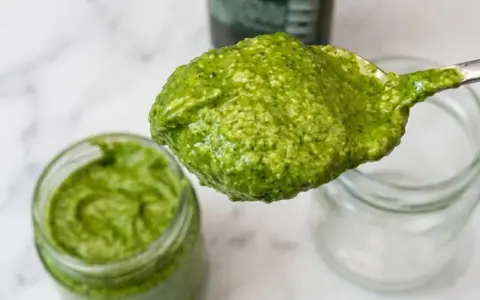 Cucchiaio pieno di pesto senza pinoli, cremoso e verde brillante, accanto a un barattolo.