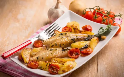 Piatto di coda di rospo al forno con patate arrosto e pomodorini. Un contorno di erbe aromatiche completa il piatto.