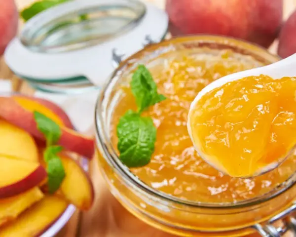 Un cucchiaio raccoglie la deliziosa marmellata di pesche da un barattolo, con pesche fresche e foglie di menta intorno.