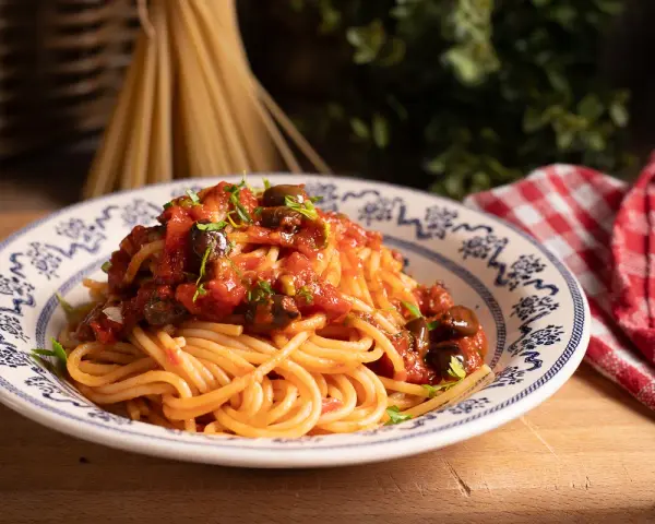 Piatto di spaghetti alla puttanesca, conditi con pomodoro, olive e capperi, guarniti con prezzemolo fresco.