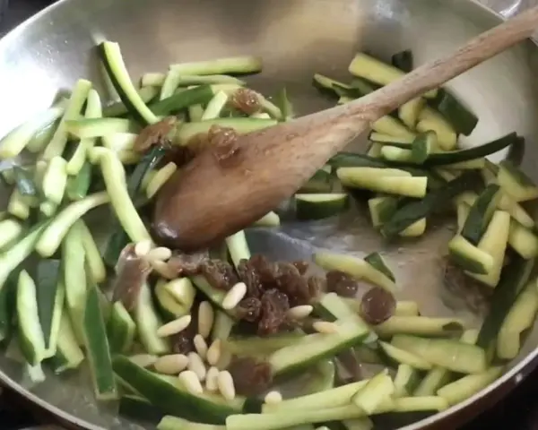 Zucchine in agrodolce veloci in padella con pinoli e uvetta, mescolate con un cucchiaio di legno.