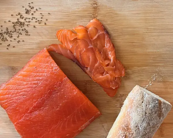 Fetta di salmone marinato, affettato e pane su tagliere di legno. Semi di cumino e sale completano la scena.