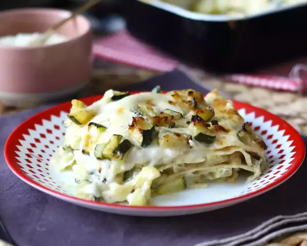 Una fetta di lasagne vegetariane con zucchine e besciamella, dorata al forno, servita su un piatto rosso e bianco.
