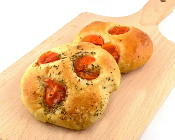 Due focaccine pugliesi dorate, condite con pomodorini e erbe aromatiche, su un tagliere di legno.