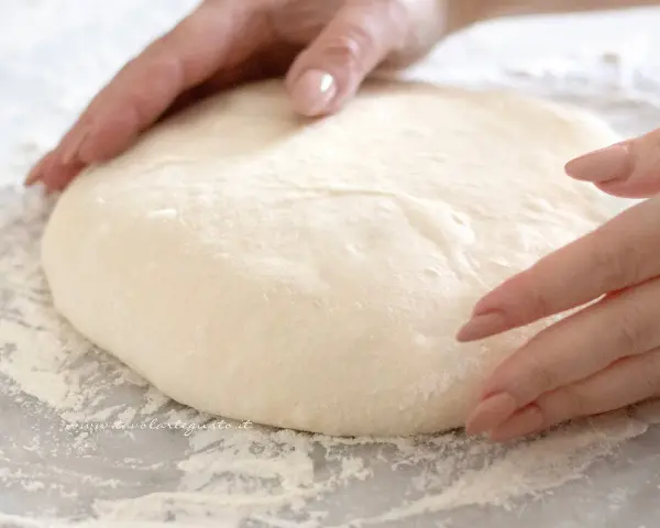 Mani che lavorano un soffice impasto pizza su un piano infarinato, pronte per essere stese.