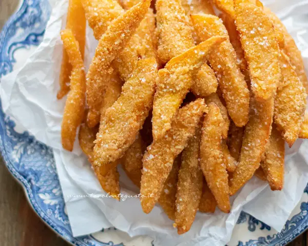 Un mucchio di bastoncini dorati di zucca fritta impanata, croccanti e salati, serviti su carta bianca in un piatto blu e bianco.