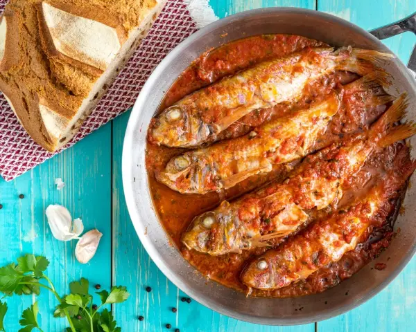 Triglie alla livornese in padella, con pane rustico, aglio e prezzemolo su tavolo blu.