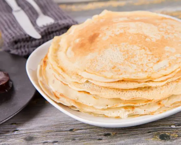 Pila di crepes senza uova, dorate e soffici, pronte per essere farcite. Accanto, un cucchiaino di marmellata.