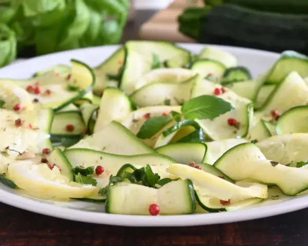 Zucchine marinate a fettine sottili, condite con erbe aromatiche e pepe rosa, su un piatto bianco.
