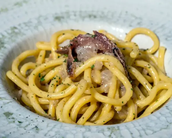 Un piatto di bigoli in salsa, con pasta spessa e condimento a base di acciughe e cipolla, guarnito con prezzemolo tritato.