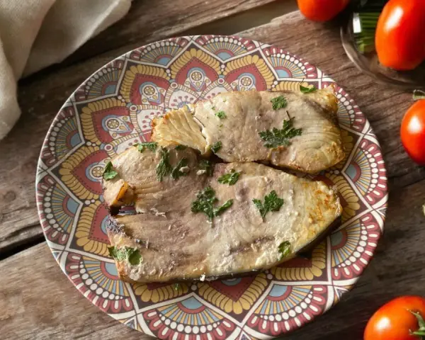 Due fette di pesce spada cotto in friggitrice ad aria, guarnite con prezzemolo, su un piatto decorato. Accanto, pomodori, limoni e zucchine.